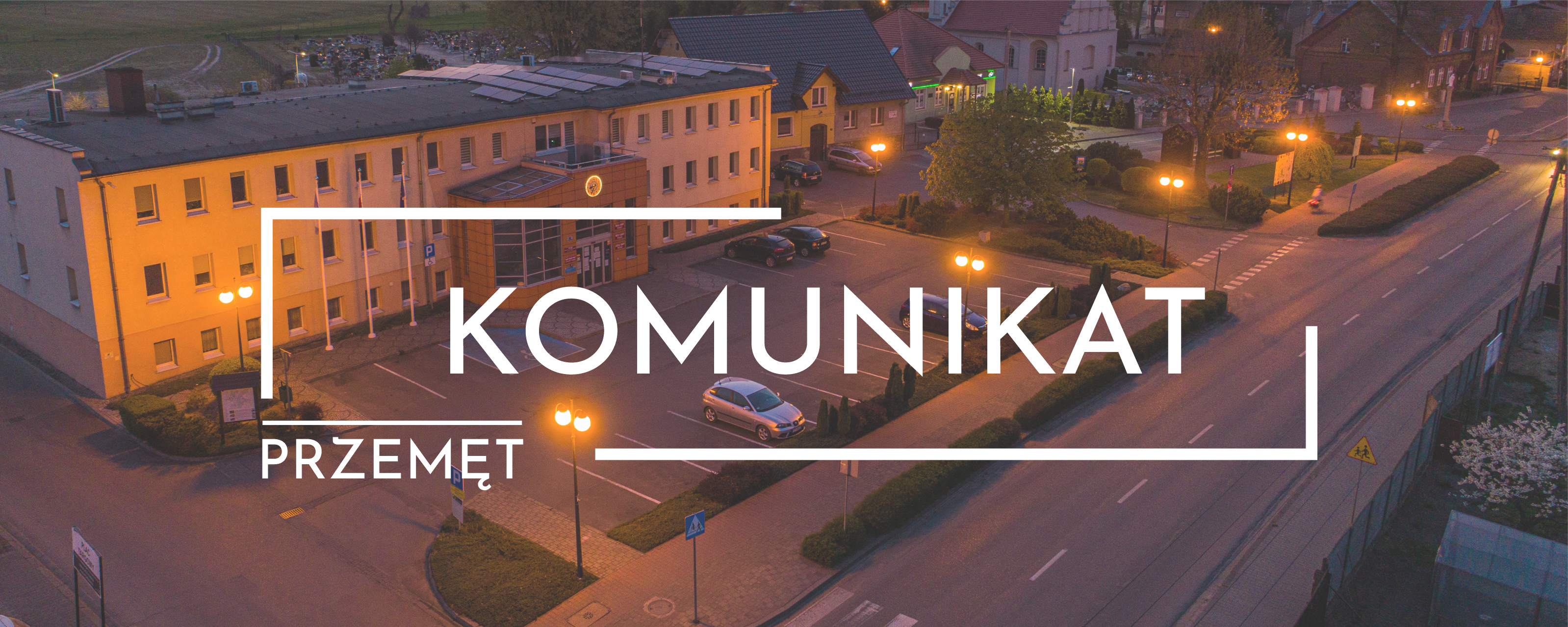 Grafika przedstawia zdjęcie urzędu gminy Przemęt i terenu wokół podczas wschodu słońca. Zdjęcie wykonano z "lotu ptaka". Na środku grafiki widnieje biały napis "komunikat".