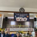 Zdjęcie przedstawia scenę, na której zawieszony jest baner, zawierający grafikę kart do gry, logo i herb gminy Przemęt oraz napis: "Przemęckie Stowarzyszenie Kopa Sportowego "Kontra" w Kluczewie". Na scenie, na stole zostały ułożone puchary, medale oraz nagrody dla zawodników. Oprócz tego na scenie umieszczono dwa roll up Gminy Przemęt. Na fotografii uchwycono także paru zawodników.