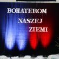 Zdjęcie przedstawia wystrój sali, w której odbywała się uroczystość. Na zdjęciu widać ściankę zrobioną z czerwonej i niebieskiej narzuty, która jest podświetlona białymi światłami. Na ściance widnieje napis "BOHATEROM NASZEJ ZIEMI"