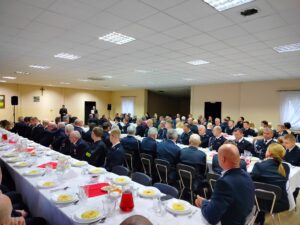 Zdjęcie przedstawia strażaków siedzących przy stołach spoglądających w kierunku sceny.