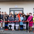 Zdjęcie przedstawia symboliczne przecięcie czerwonej wstęgi przez zaproszonych gości, uczestników turnieju i gospodarzy jako znak otwarcia stadionu.
