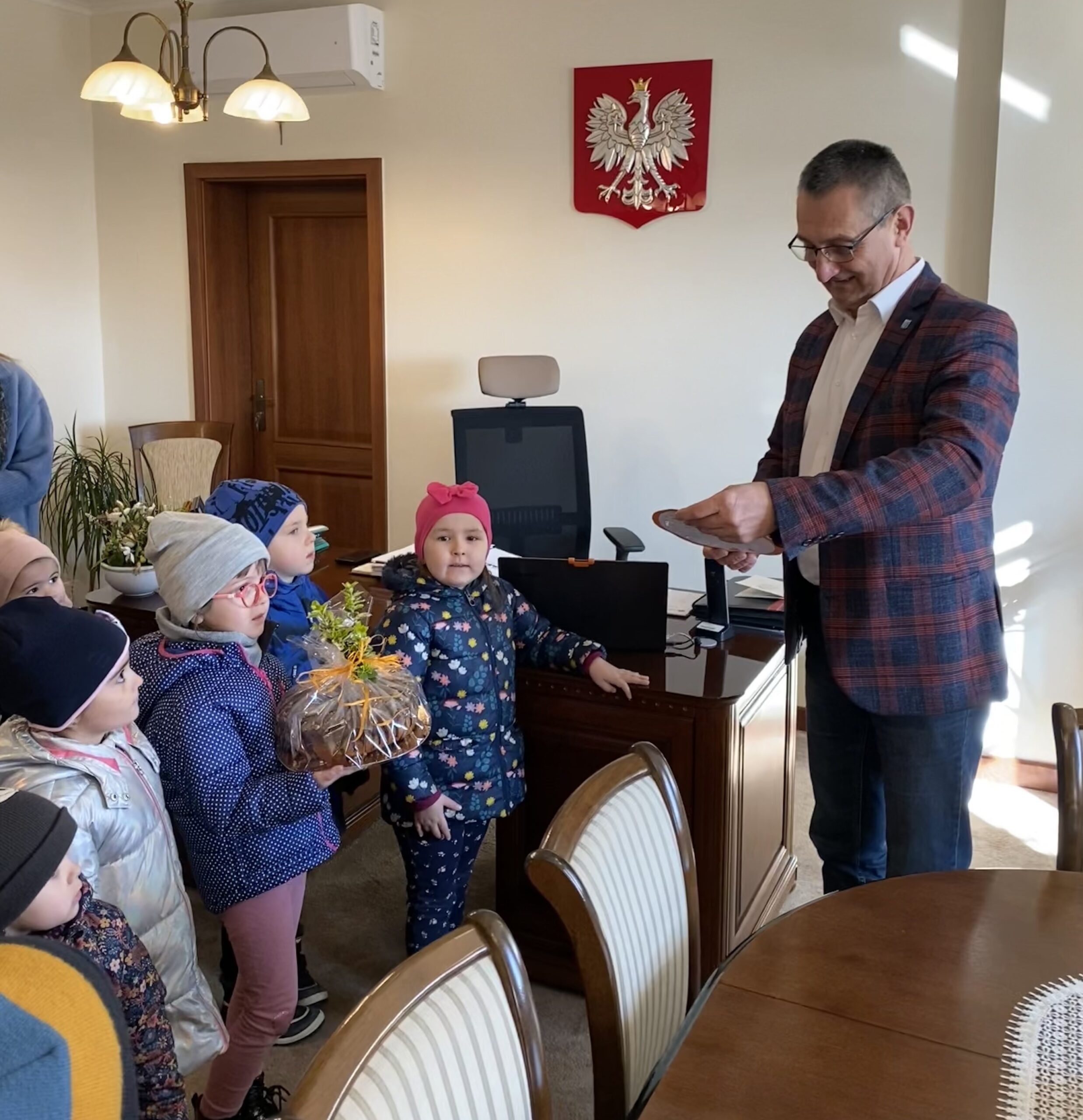 Zdjęcie przedstawia Wójta, który odbiera od dzieci podarunki. Z uśmiechem na twarzy czyta życzenia wielkanocne od przedszkolaków.