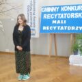 Zdjęcie przedstawia dziewczynę podczas recytacji swojego wiersza. W tle za nią widać tablicę, na której widnieje biały napis na niebieskim tle: "Gminny konkurs recytatorski. Mali recytatorzy".