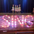 Zdjęcie przedstawia scenę, na przodzie której widnieje duży różowy napis w kropki: "SING", który jest podświetlony. Na scenie występuje czwórka dziewczynek, które ubrane są w różowe spódniczki i białe bluzki, a na głowach mają ubrane królicze uszy.