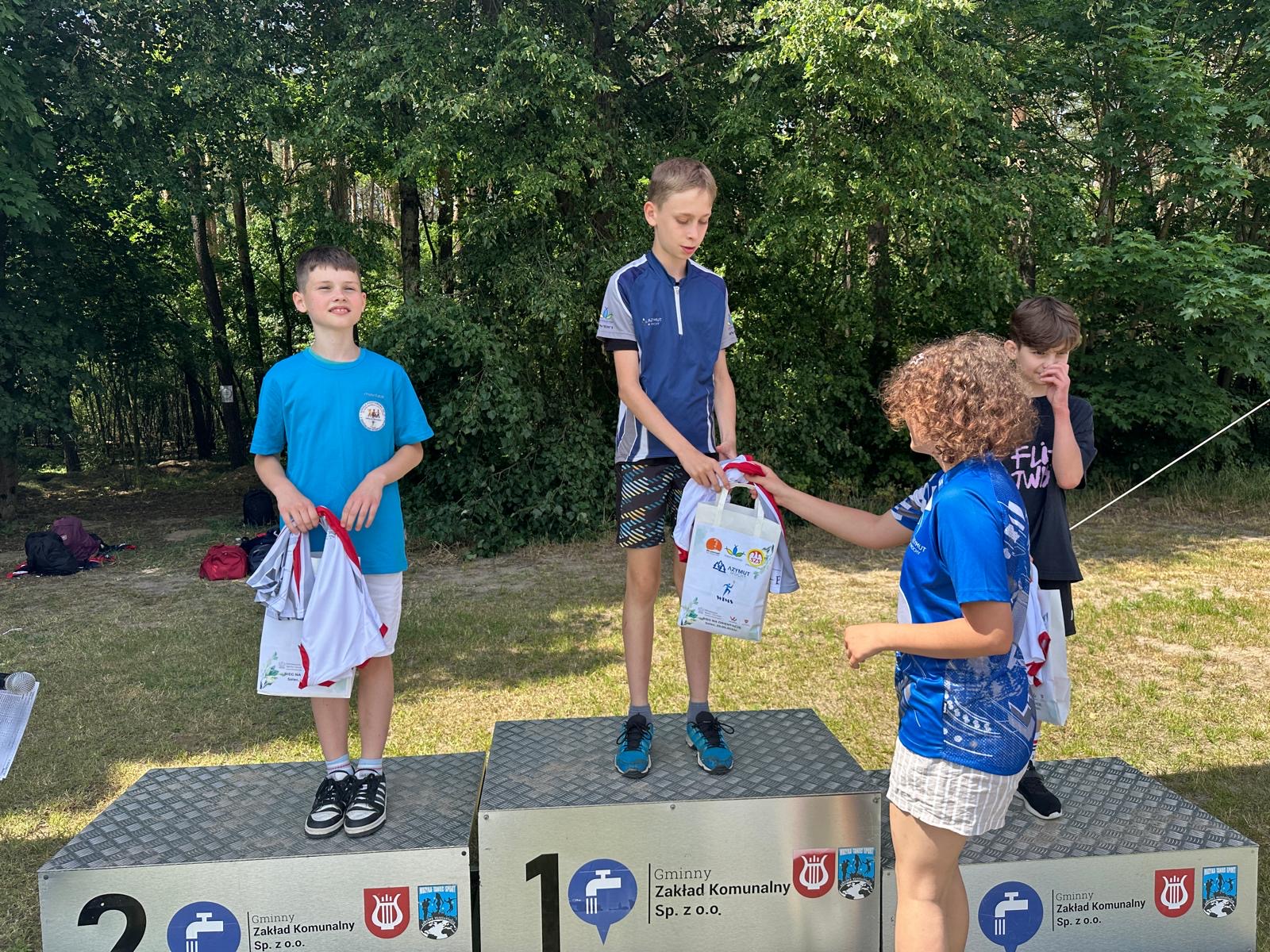 Zdjęcie przedstawia trzech chłopaków, którzy stoją na podium. Kobieta wręcza nagrodę, temu który stoi na najwyższym podium.