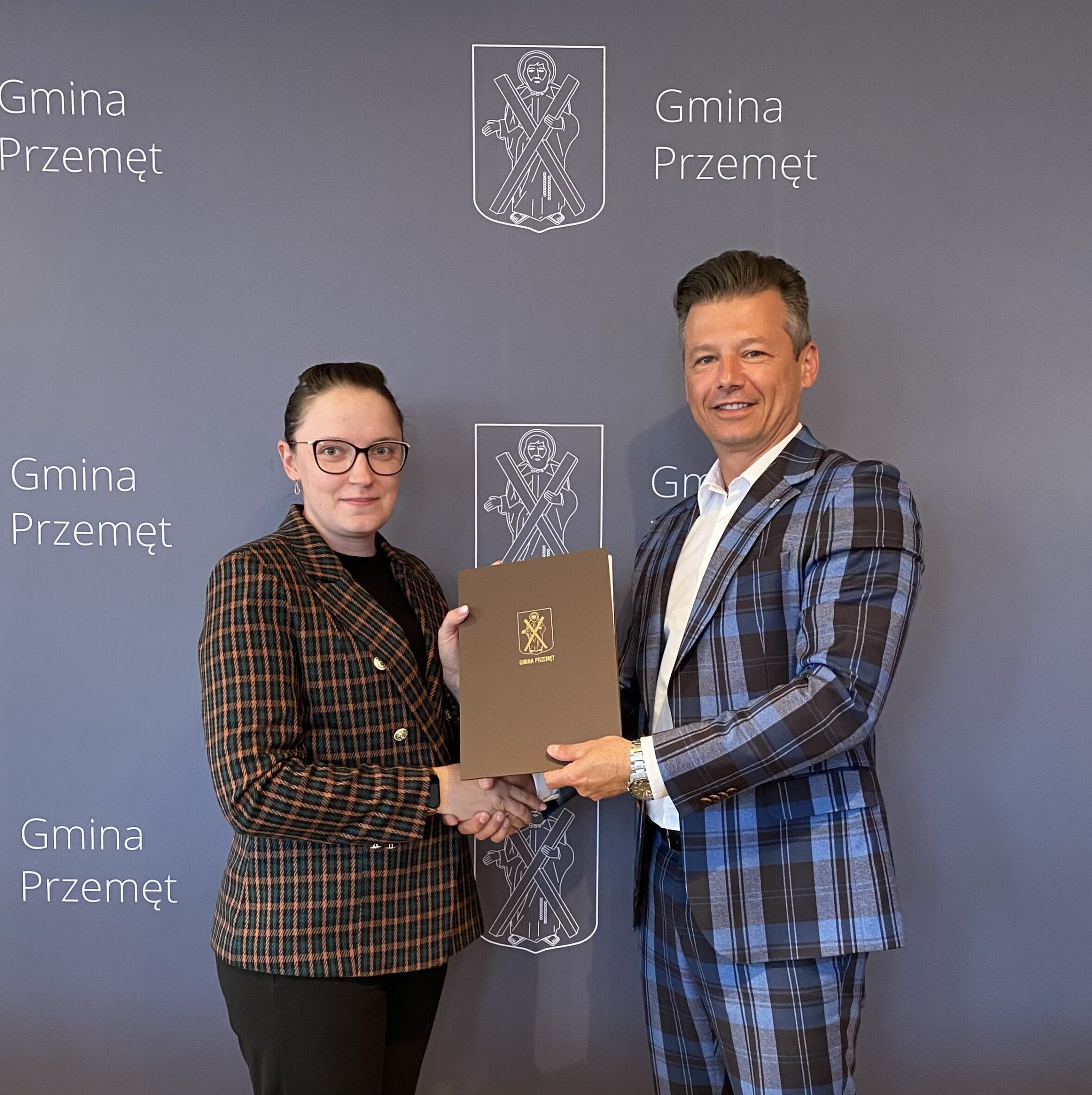 Na zdjęciu zastępca wójta Gminy Przemęt Waldemar Kalitka wraz z pełnomocnikiem firmy Panią Katarzyną Mocek. Wyżej wymienieni podają sobie dłonie, a w rękach trzymają podpisaną umowę w teczce Gminy Przemęt. W tle ścianka "Gmina Przemęt".