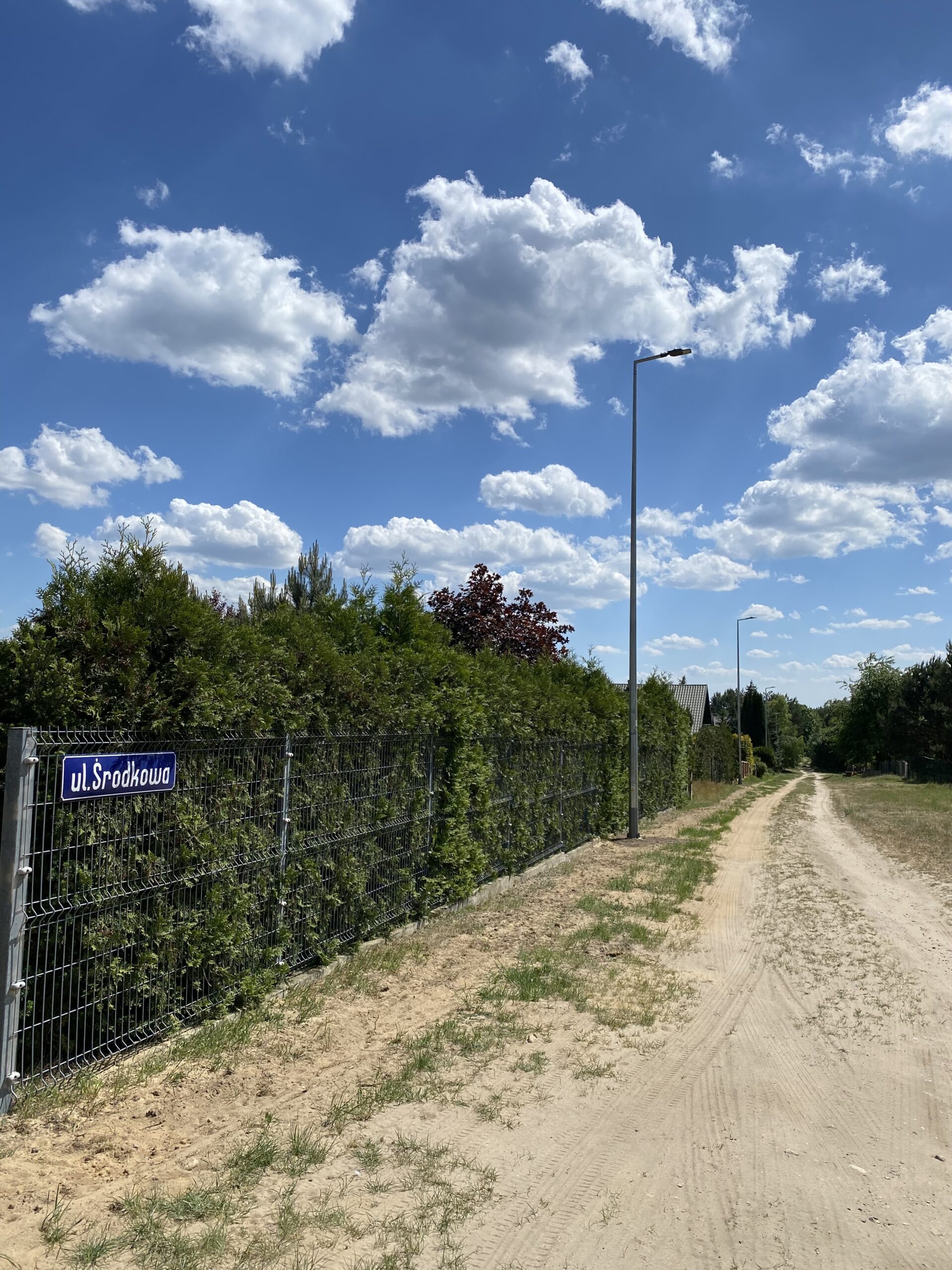 Zdjęcie przedstawia oświetlenie na ulicy Środkowej. Na zdjęciu droga piaszczysta, wokół krzewy i ogrodzenie, na której znajduję się tabliczka z napisem "Środkowa".