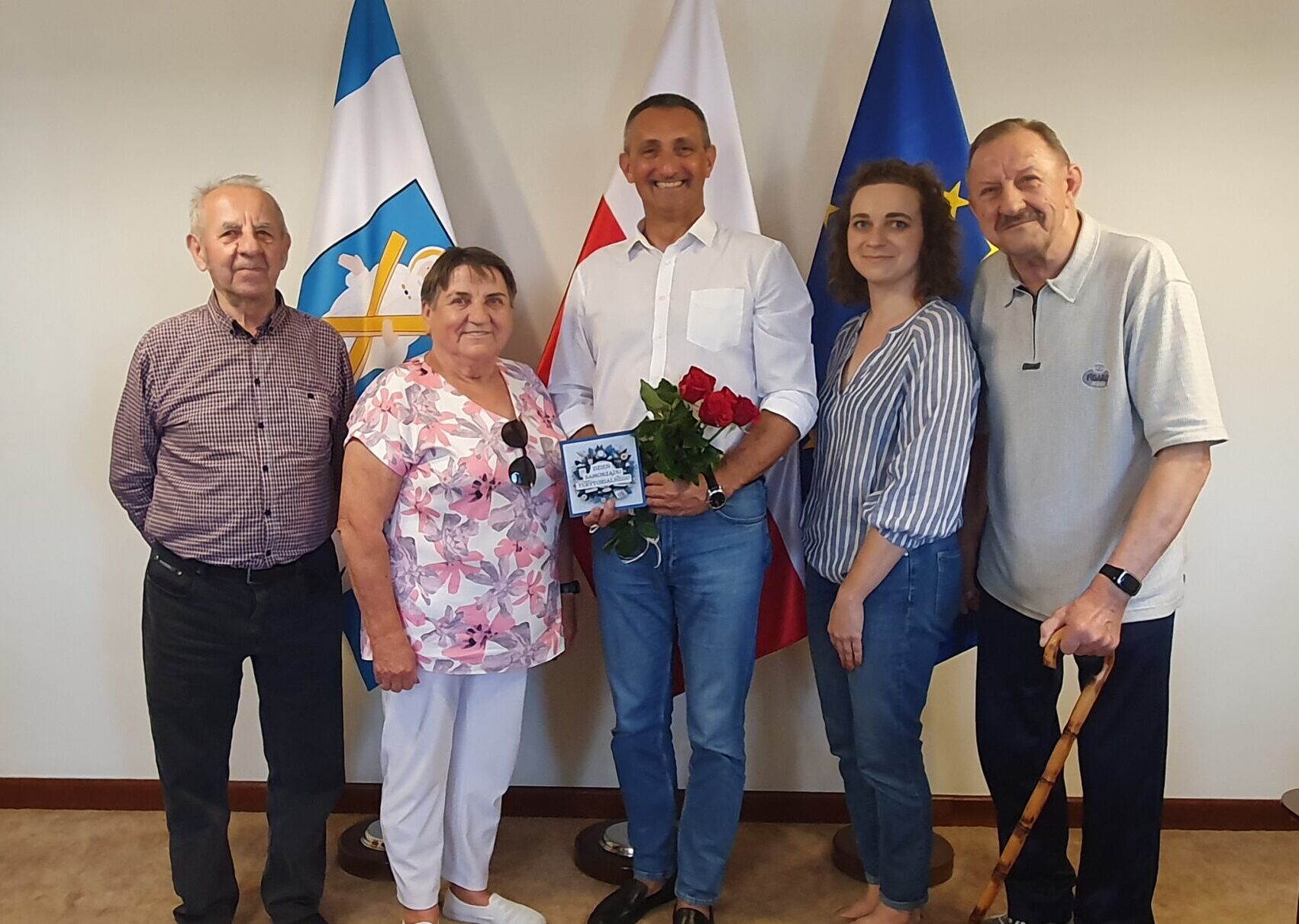 Na zdjęciu seniorzy z DDP w Błotnicy wraz z wójtem. Wójt trzyma w ręce bukiet czerwonych róż oraz ozdobną kartkę z podziękowaniami. Wszyscy pozują na tle flag w biurze wójta.