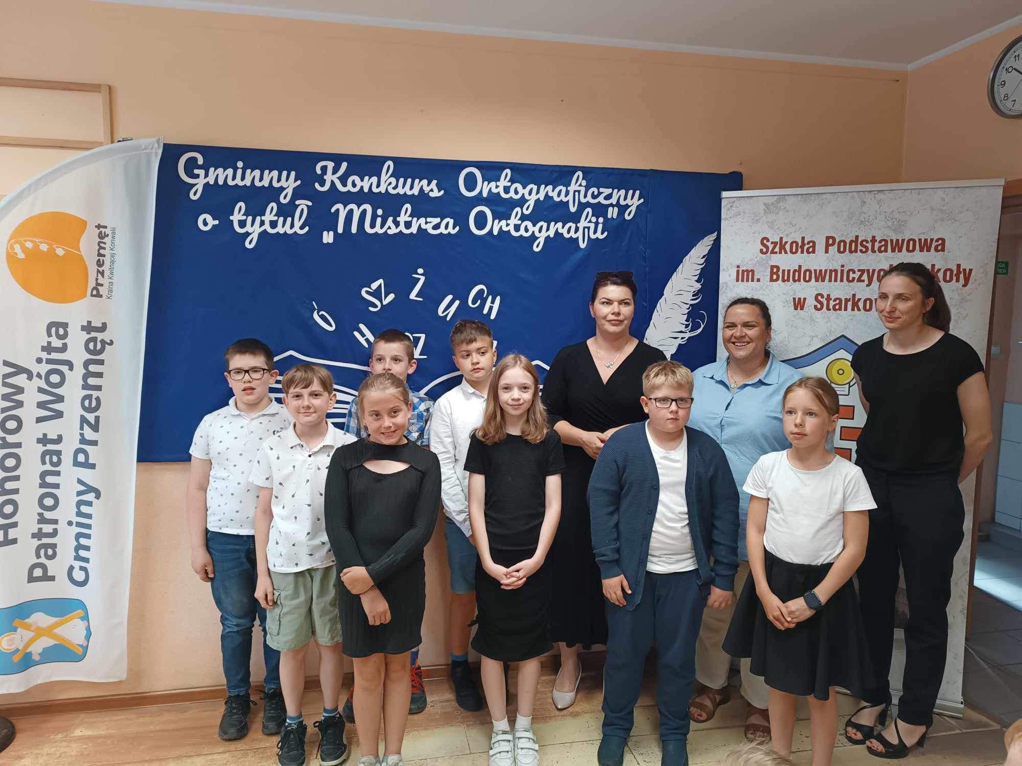 Wspólne zdjęcie uczestników konkursu wraz z przewodniczącą rady gminy Przemęt Panią Moniką Maćkowiak oraz Panią Dyrektor Lidią Ławecką. Wszyscy pozują na tle tablicy, na której widnieje napis: "Gminny Konkurs Ortograficzny o tytuł mistrza Ortografii". Na zdjęciu znajdują się też windery honorowy patronat wójta Gminy Przemęt oraz roll up szkoły w Starkowie.