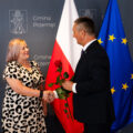 Na zdjęciu uchwycono moment uścisku dłoni wójta wraz z byłą Panią sołtys Solca Nowego. Wójt trzyma w drugiej ręce czerwoną różę. W tle ścianka oraz flagi.