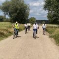Grupa rowerzystów jedzie drogą polną. W tle drzewa i pola.