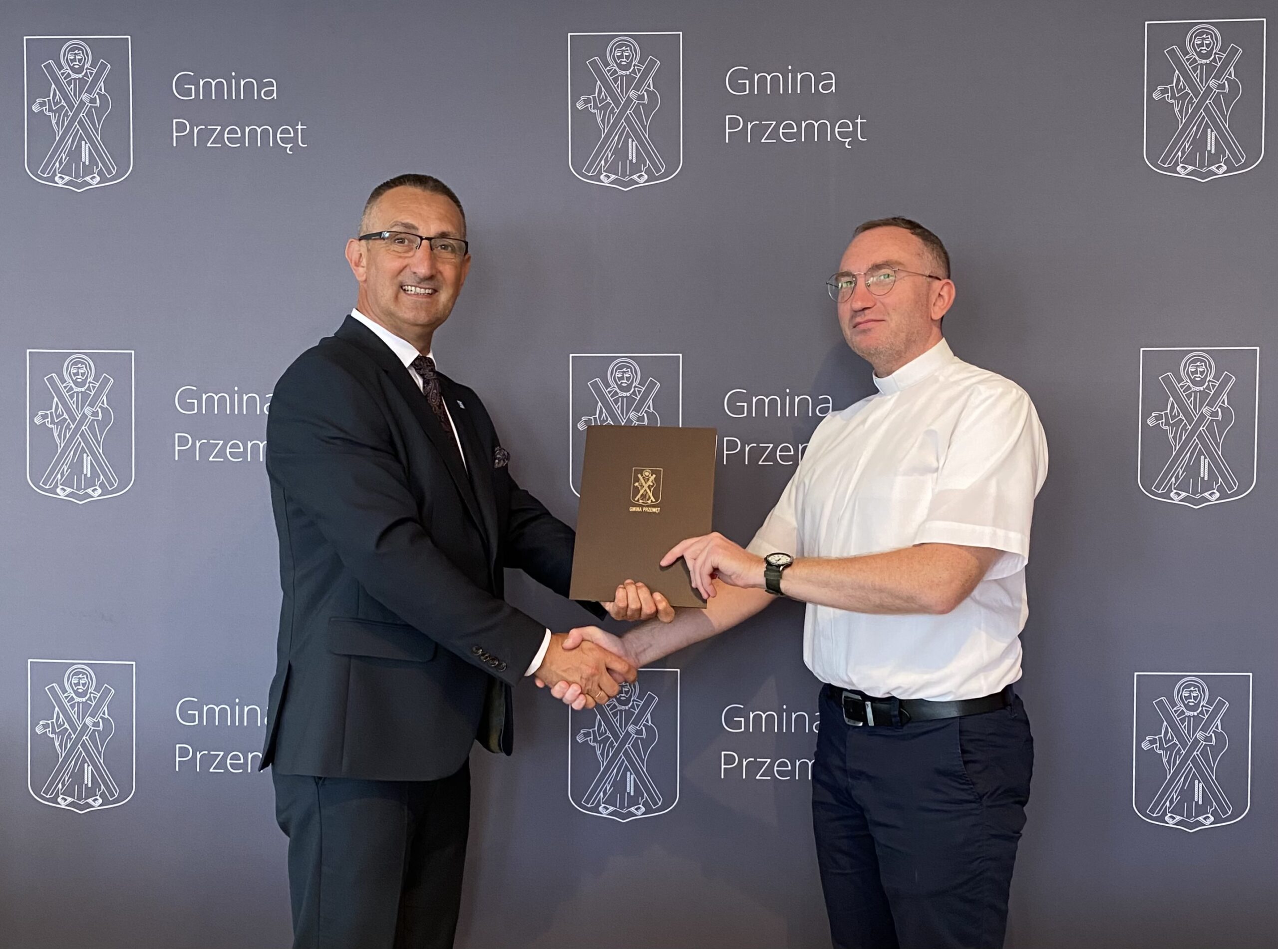 Zdjęcie przedstawia Wójta Gminy Przemęt wraz z księdzem proboszczem z parafii Rzymskokatolickiej pod wezwaniem św. Barbary w Buczu. Osoby ww. podają sobie dłoń na tle ścianki Gmina Przemęt.