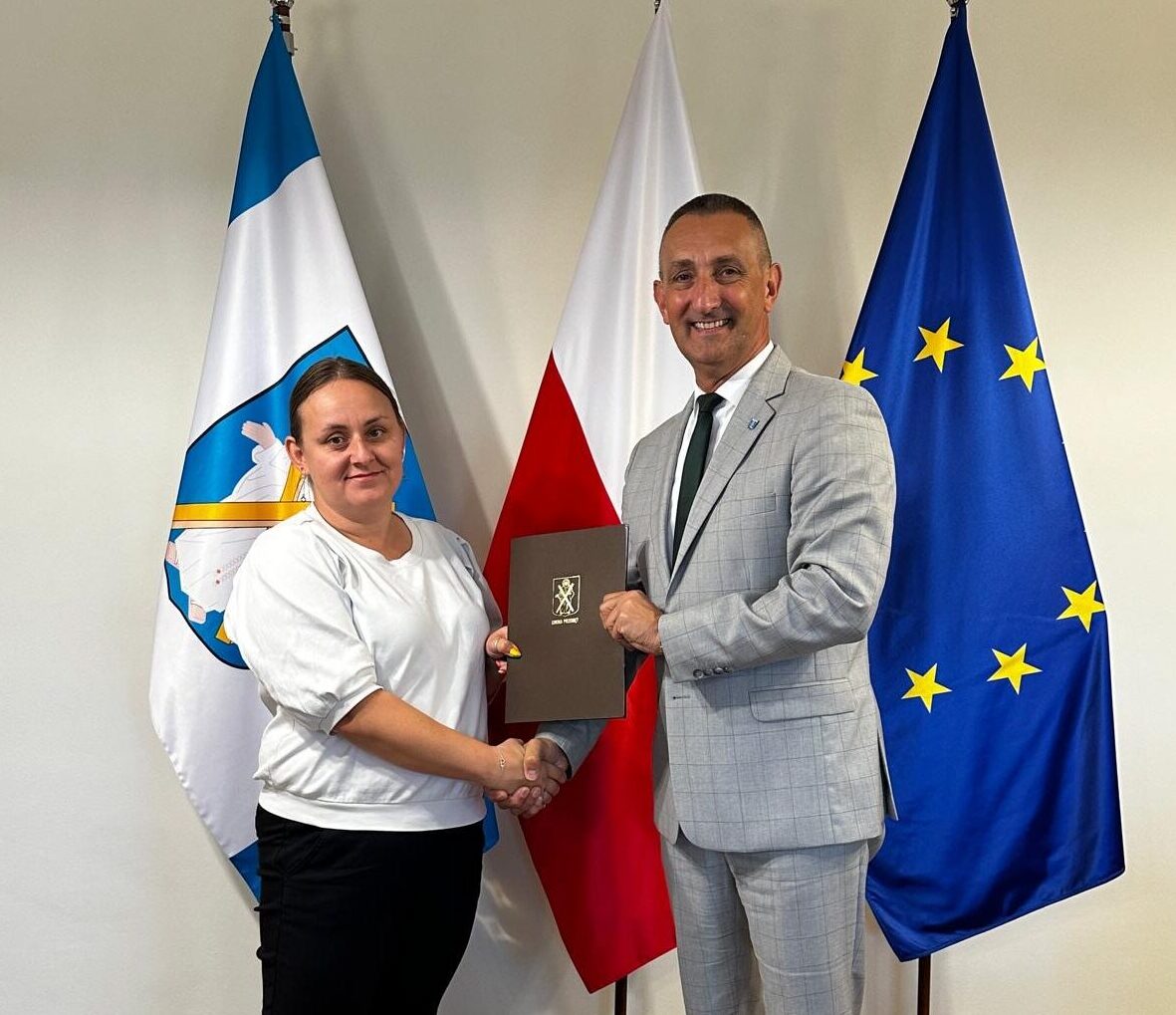 Zdjęcie przedstawia Wójta Gminy Przemęt wraz z Prezesem Zarządu Agnieszką Nowak. Osoby ww. podają sobie dłoń oraz trzymają teczkę z herbem Gminy na tle flag: Polski, Europejskiej oraz flagi z herbem św. Andrzeja z krzyżem.