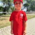 Na zdjęciu uśmiechnięta dziewczynka ubrana w czerwoną koszulkę i czapkę z daszkiem. Ubrania maja oznakowania OSP Przemęt.