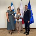 Na zdjęciu wójt, kierownik oświaty oraz dyrektor Edyta Kaczmarek. Wszyscy pozują w gabinecie na tle flag: polskiej, europejskiej oraz flagi z herbem gminy. Pani Edyta trzyma róże oraz akt powierzenia.