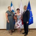 Na zdjęciu wójt, kierownik oświaty oraz dyrektor Barbara Matysiak. Wszyscy pozują w gabinecie na tle flag: polskiej, europejskiej oraz flagi z herbem gminy. Pani Barbara trzyma róże oraz akt powierzenia.