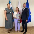 Na zdjęciu wójt, kierownik oświaty oraz dyrektor Alina Piotrowska. Wszyscy pozują w gabinecie na tle flag: polskiej, europejskiej oraz flagi z herbem gminy. Pani Alina trzyma róże oraz akt powierzenia.