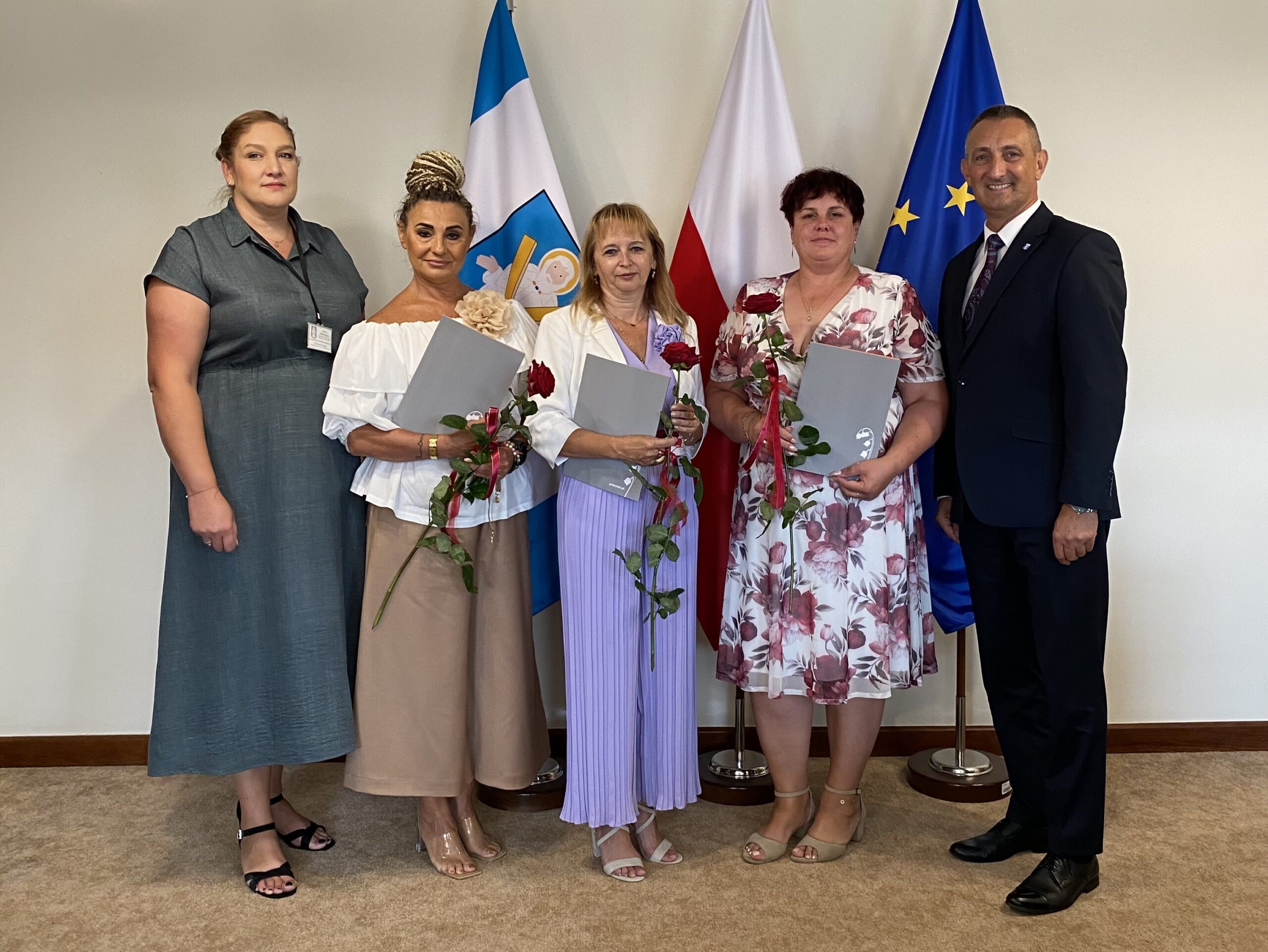 Na zdjęciu wójt, kierownik oświaty oraz dyrektorki. Wszyscy pozują w gabinecie na tle flag: polskiej, europejskiej oraz flagi z herbem gminy. Dyrektorki trzymają róże oraz akty powierzenia.