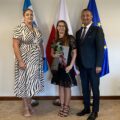 Na zdjęciu wójt, kierownik oświaty oraz Pani Agnieszka Rimke. Wszyscy pozują w gabinecie na tle flag: polskiej, europejskiej oraz flagi z herbem gminy. Pani Agnieszka trzyma róże oraz awans zawodowy.