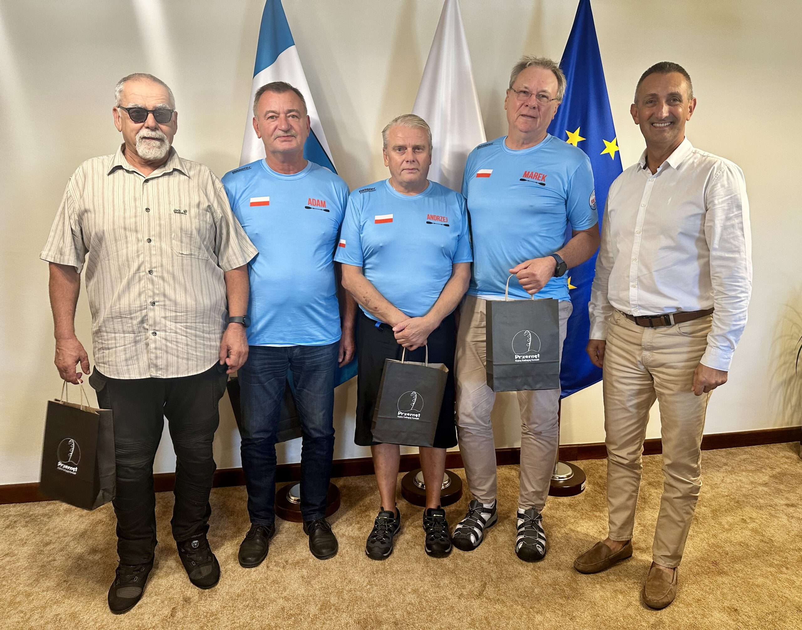 Grupowe zdjęcie, na którym jest wójt wraz z uczestnikami wyprawy, którzy trzymają upominki. Wyżej wymienieni pozują w biurze na tle flag: polskiej, europejskiej oraz flagi z herbem gminy Przemęt.