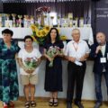 Na zdjęciu Pani Patrycja Pabich, Pan Zdzisław Kaźmierczak, Pani Danuta Buchwald, Pani Jolanta Skoracka oraz osoba, które prowadzi wydarzenie. Pani Patrycja oraz Pani Jolanta trzymają bukiety kwiatów. Wszyscy pozują na tle sceny, na której znajdują się nagrody oraz puchary dla uczestników turnieju.