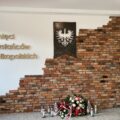 Zdjęcie przedstawia nową ścianę upamiętniającą bohaterów Powstania Wielkopolskiego. Połowa ściany została pokryta białą farbą, a druga połowa wyłożona cegłą. Na ścianie zawieszono godło Polski oraz zamieszczono złoty napis: "Pamięci Powstańców Wielkopolskich". Pod ścianką znajdują się wieńce oraz znicze.