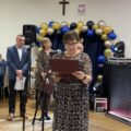 Kobieta czyta życzenia przez mikrofon, w tle inne osoby.