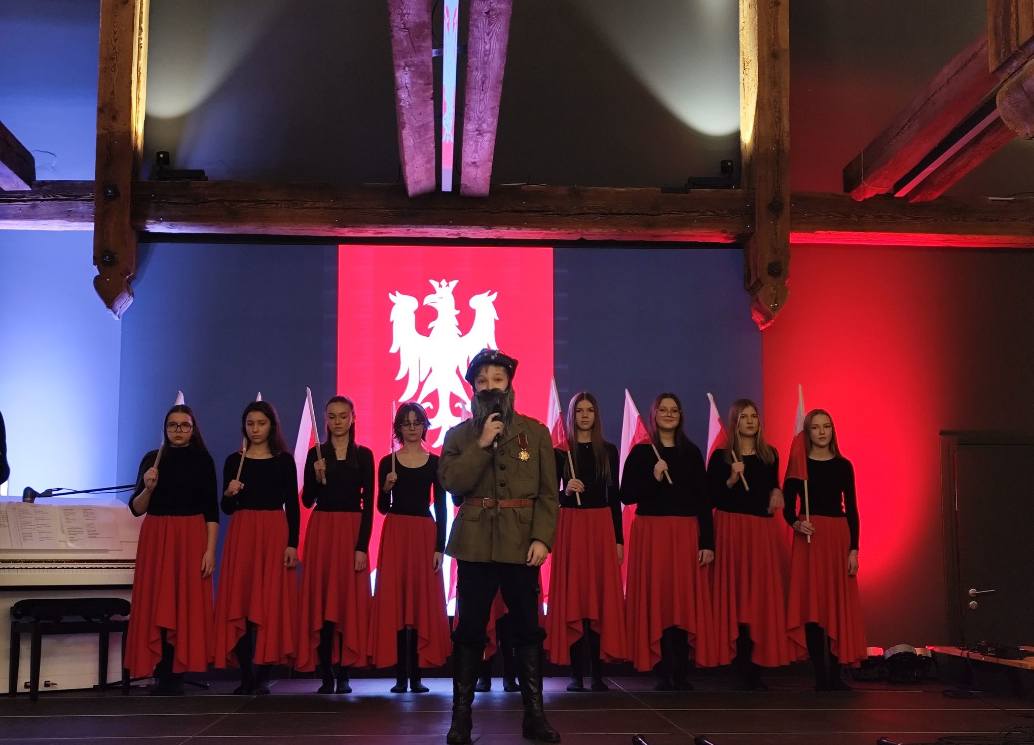 Zdjęcie przedstawia występ artystyczny w wykonaniu dzieci ze Szkoły Podstawowej im. Ignacego hr. Mielżyńskiego w Chobienicach. Dziewczyny ubrane są w długie czerwone spódnice, a chłopak stojący na środku przebrany jest za żołnierza. W tle wyświetlona została flaga powstania wielkopolskiego.