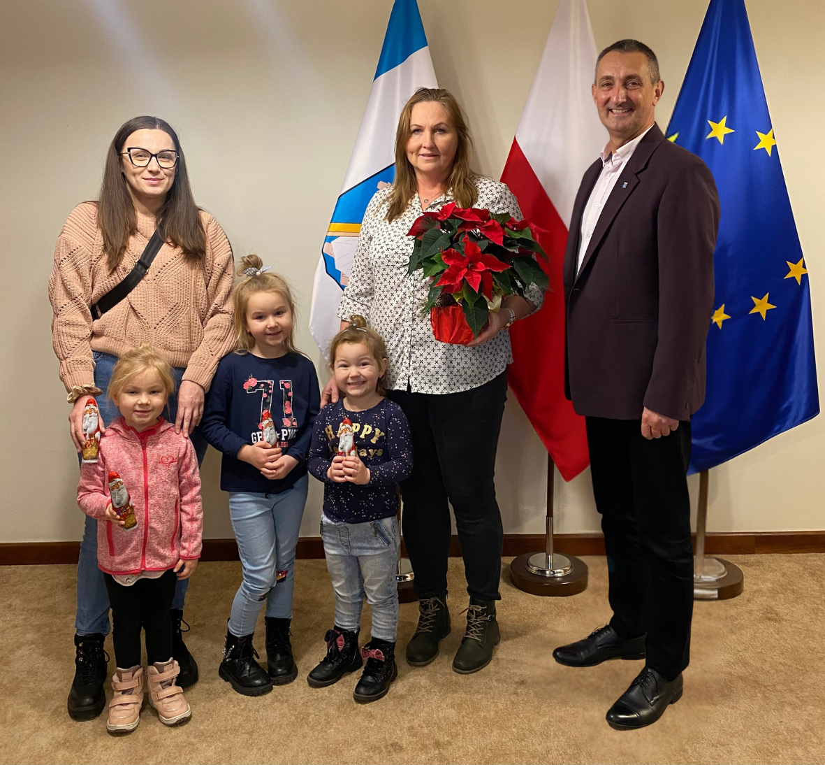 Na zdjęciu wójt Janusz Frąckowiak, dyrektor przedszkola w Przemęcie Pani Elżbieta Grygiel-Tórz, nauczycielka z przedszkola oraz trzy dziewczynki, które trzymają czekoladowe gwiazdorki. Pani dyrektor trzyma kwiat gwiazdę betlejemską. Wszyscy pozują na tle flag w gabinecie wójta.