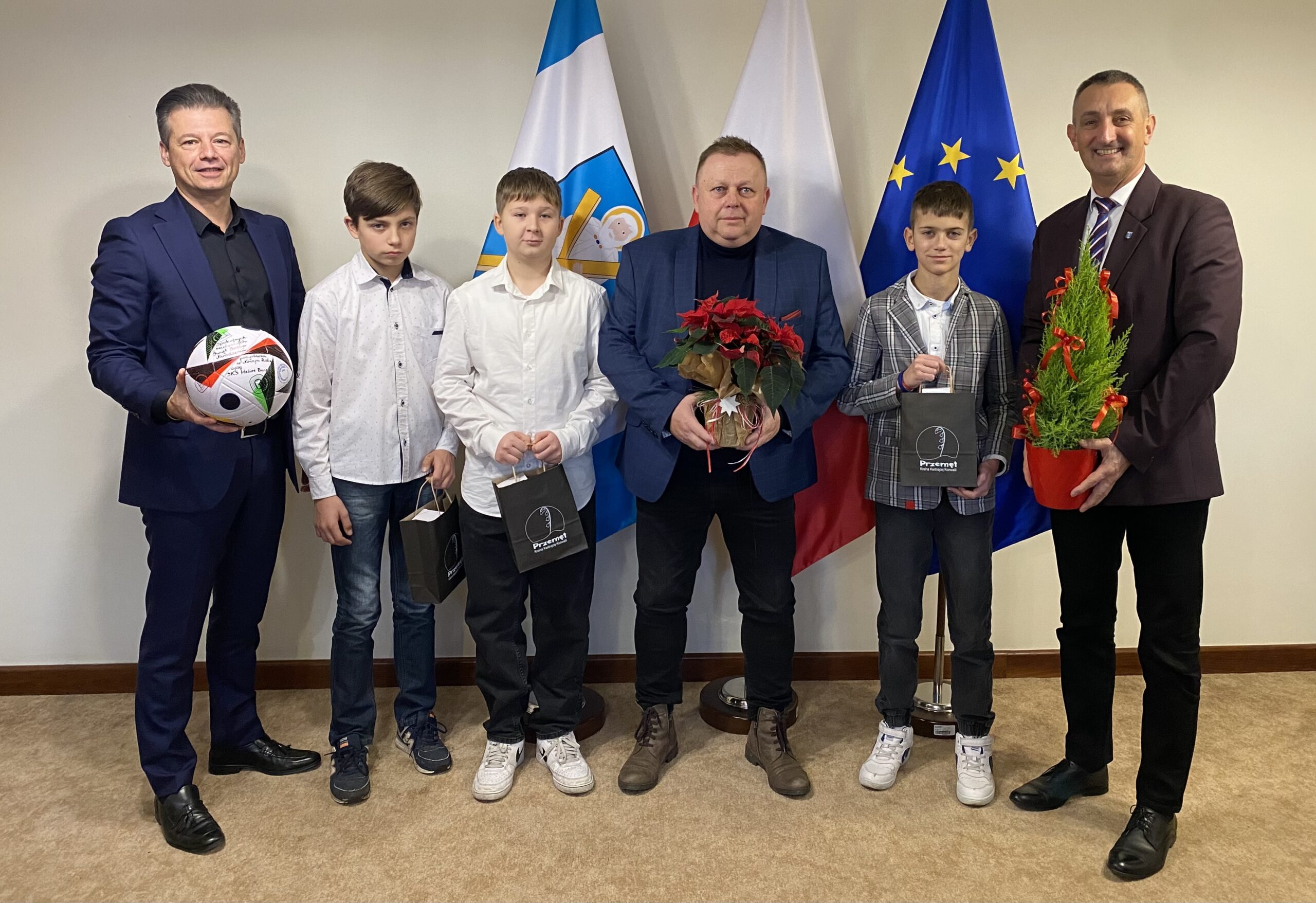 Na zdjęciu wójt Janusz Frąckowiak, który trzyma choinkę świąteczną, zastępca wójta Waldemar Kalitka, który trzyma podpisaną przez zawodników klubu piłkę, prezes klubu Robert Muller, który trzyma kwiat gwiazdę betlejemską oraz zawodnicy klubu z upominkami. Wszyscy pozują na tle flag w gabinecie.