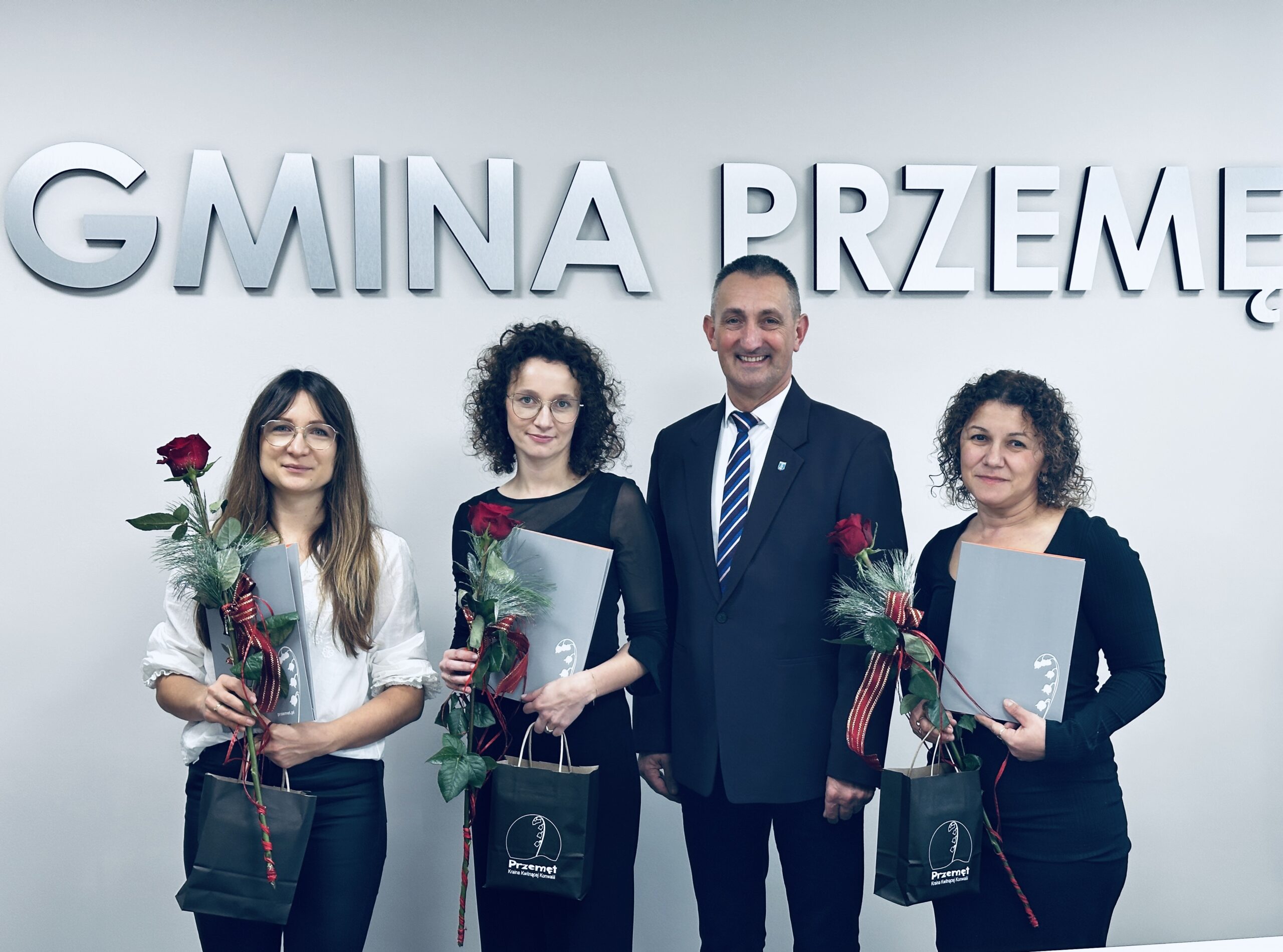 Na zdjęciu wójt Janusz Frąckowiak wraz z nauczycielkami: Joanną Bednarczyk, Pauliną Przybyłą i Bernadetą Spławską. Panie trzymają teczki, w których są akty mianowania oraz róże i drobne upominki. Wszyscy pozują na tle srebrnego napisu Gmina Przemęt.