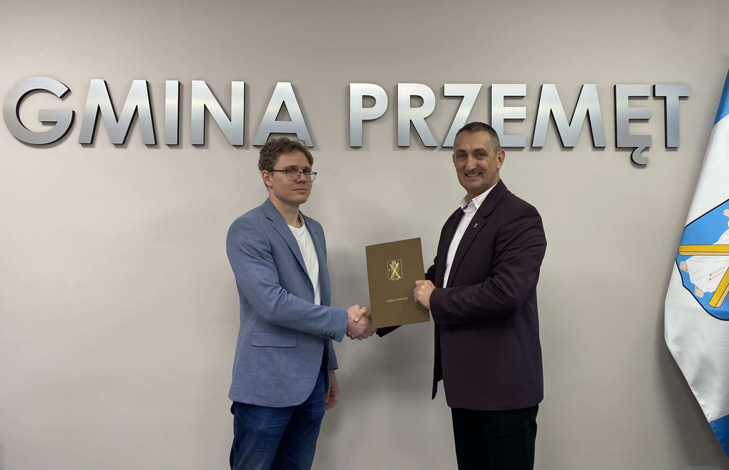 Na zdjęciu wójt wraz z panem Mateuszem Sitą. Wyżej wymienione osoby podają sobie dłoń i trzymają teczkę, w której znajduje się podpisana umowa. W tle srebrny napis "Gmina Przemęt" oraz flaga z herbem gminy.