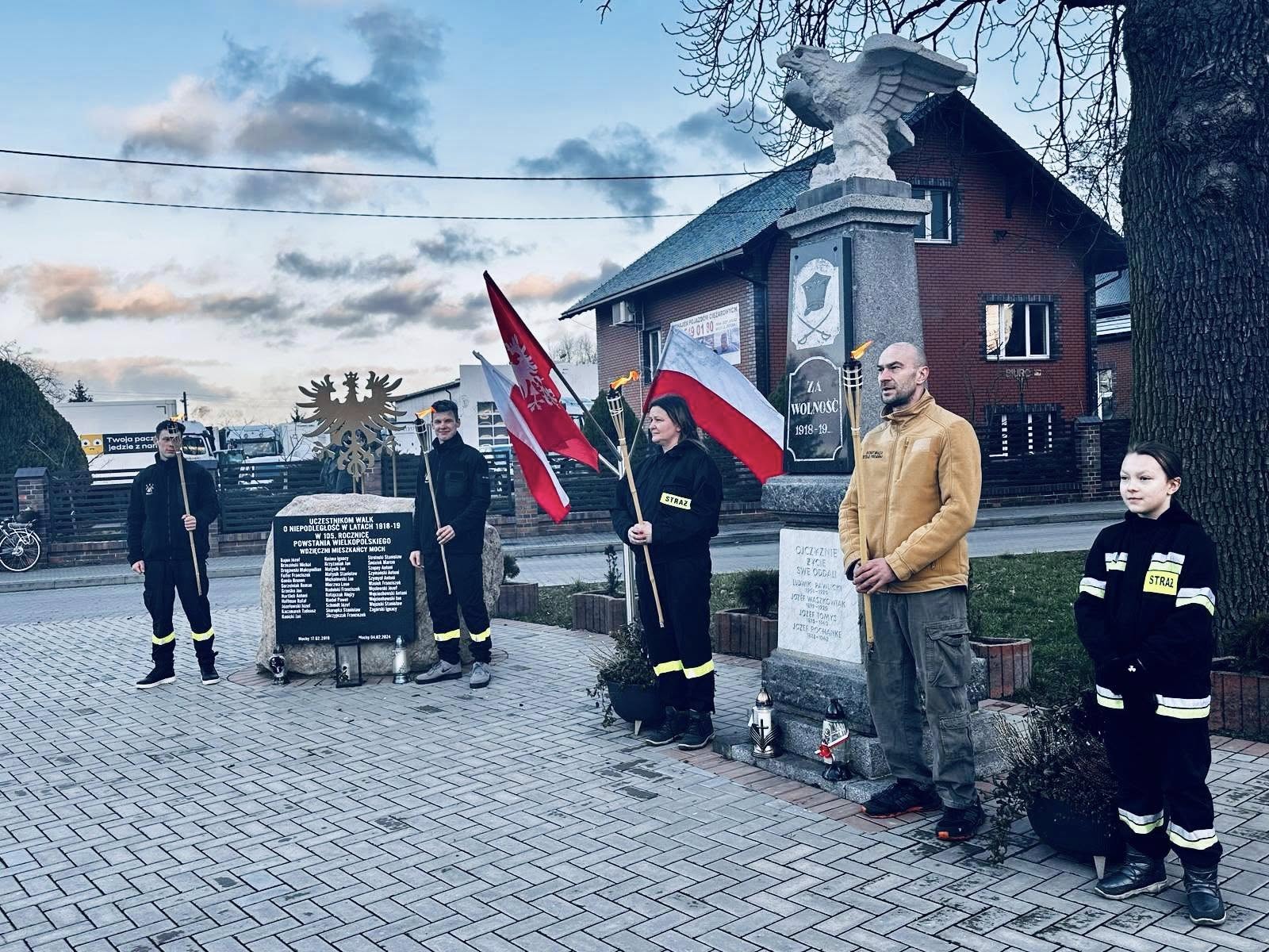 Na zdjęciu osoby należące do OSP Mochy stoją przy pomniku Powstańców Wielkopolskich i przy tablicy pamiątkowej w Mochach.