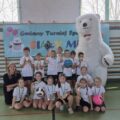 Grupowe zdjęcie zawodników z Kaszczoru wraz z opiekunami i osobą przebraną za białego misia. Zawodnicy mają założone medale, trzymają dyplom, puchar oraz nagrody w postaci piłek. W tle plakat z napisem Gminny Turniej Sportowy Biały Miś oraz winder Honorowy Patronat Wójta Gminy Przemęt.