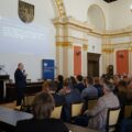 Na zdjęciu osoba przemawia do reszty osób siedzących w sali. Na projektorze wyświetlana jest informacja związana z zasadami otrzymywania i rozliczania dotacji celowych.