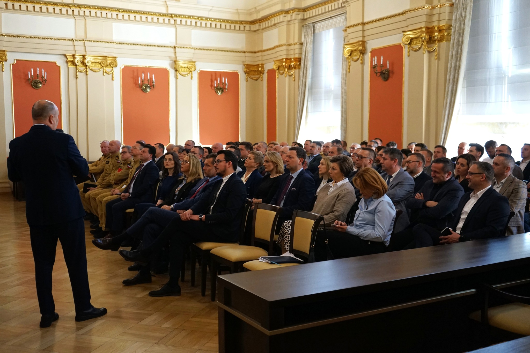 Na zdjęciu osoby obecne na konferencji. Wszyscy znajdują się w sali. Jedna osoba przemawia do reszty osób.