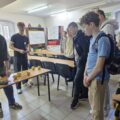 Po lewej stronie dwóch chłopaków pokazuje eksperyment grupie nastolatków, która stoi z prawej strony.