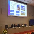 Na zdjęciu kobieta, która przemawia podczas konferencji. Na fotografii także czwórka innych osób. Na projektorze wyświetlana jest prezentacja o tytule: "Ochrona zabytków i budownictwa tradycyjnego".