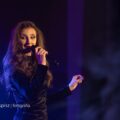 Na zdjęciu Ksenia Shaushyshvili podczas występu na scenie. Kobieta ma ubraną długą, czarną, wieczorową suknię.