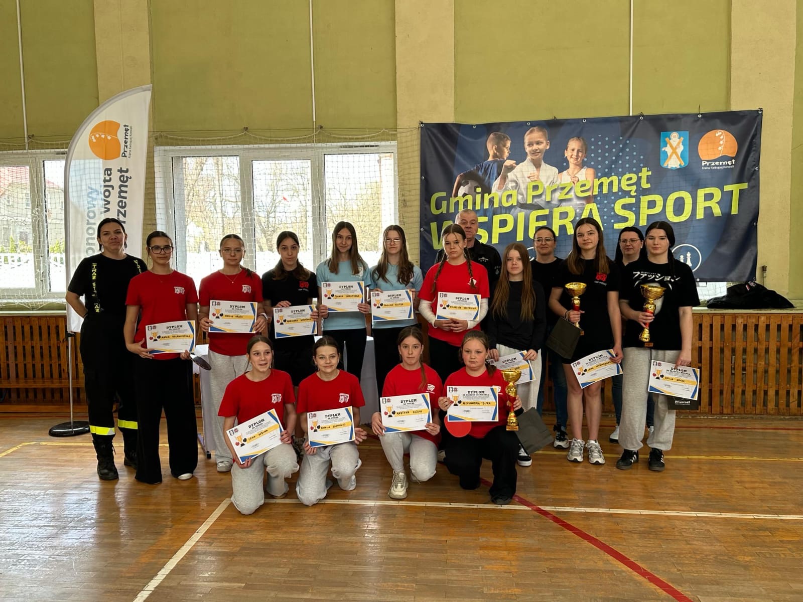 Zdjęcie grupowe dziewcząt. Każda dziewczyna pozuje z dyplomem, a zwyciężczynie z dyplomami, pucharami i upominkami. Na fotografii także Wiceprezes Zarządu Oddziału Gminnego ZOSP RP w Przemęcie dh Marian Skibiński, Pani Julita Nitschke oraz dwie inne kobiety. W tle baner "Gmina Przemęt wspiera sport".