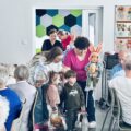 Fotografia ukazuje moment wejścia do sali dzieci ze żłobka wraz z opiekunkami. Dzieci przyniosły ze sobą koszyczki oraz ozdoby wielkanocne w postaci papierowych zajączków.
