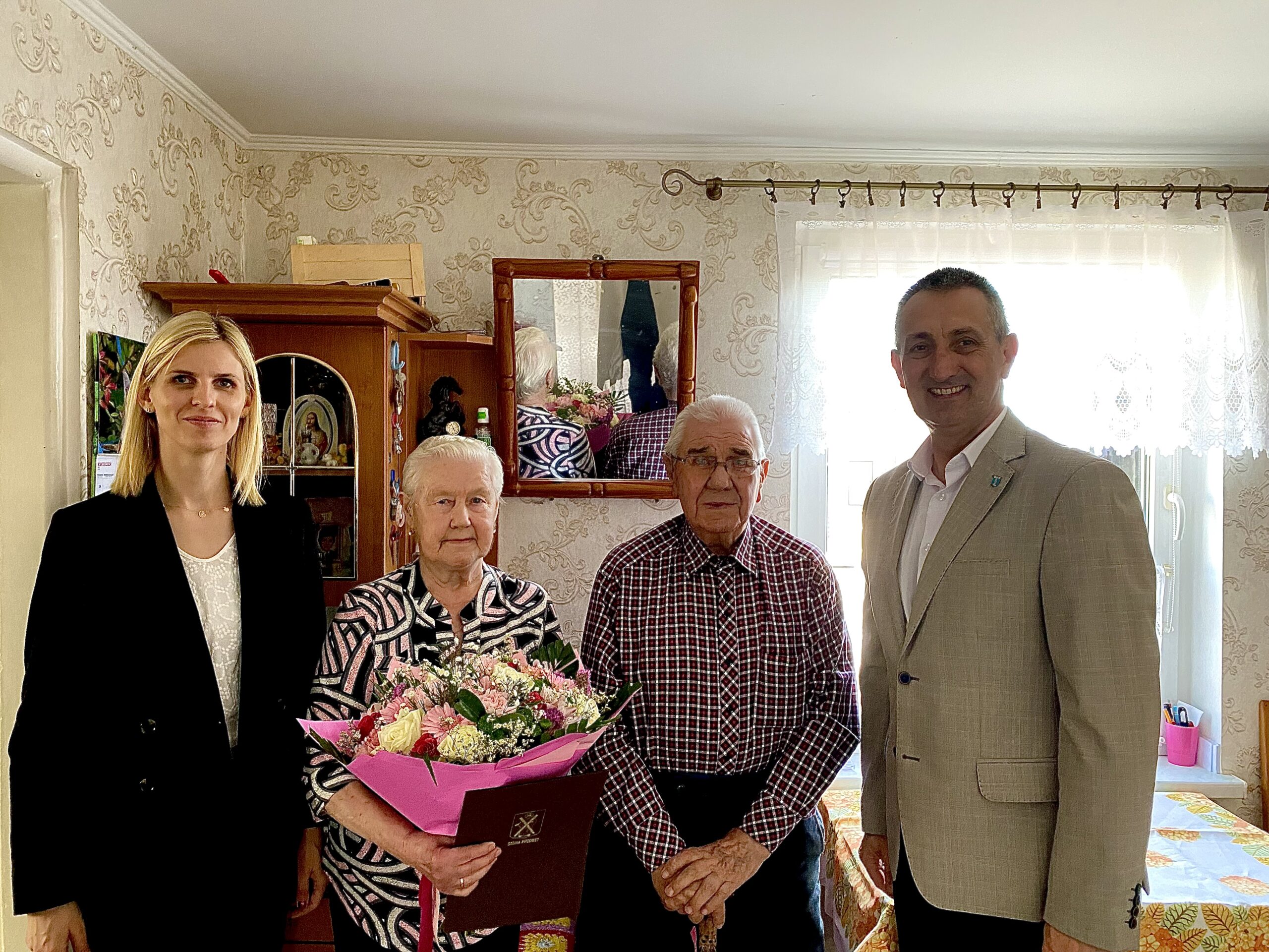 Na zdjęciu wójt, Pani Irena - zastępca kierownika, jubilatka wraz z mężem. Jubilatka trzyma bukiet kwiatów i list gratulacyjny.