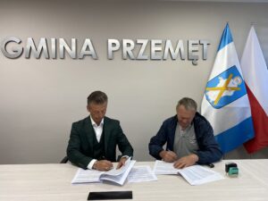 Zdjęcie przedstawia zastępcę wójta Waldemara Kalitkę oraz przedstawiciela firmy Zygmunta Marszałka podczas podpisywania umowy. W tle srebrny napis: "GMINA PRZEMĘT" oraz flaga Polski i flaga z herbem gminy Przemęt.