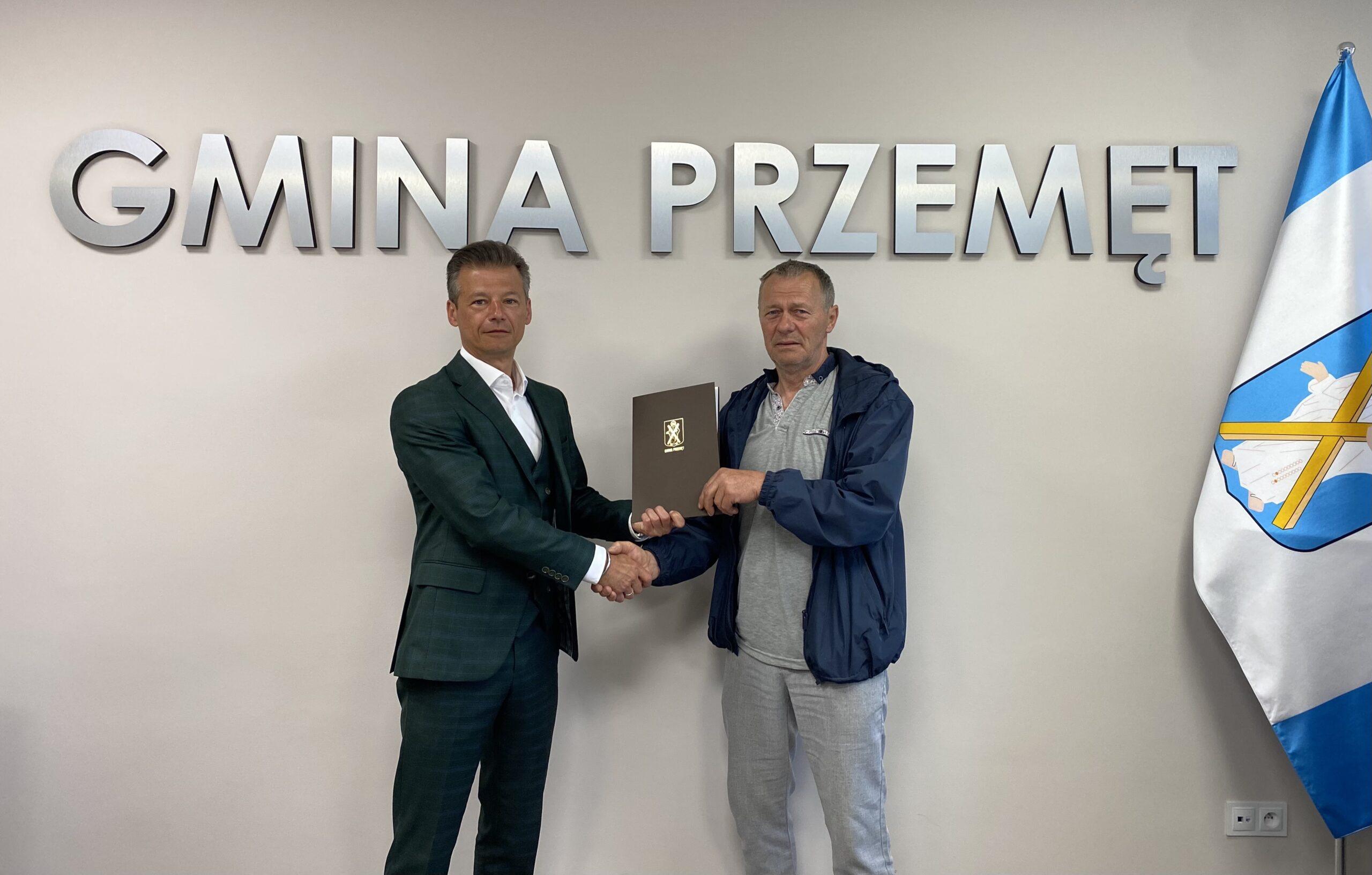 Zdjęcie przedstawia zastępcę wójta Waldemara Kalitkę oraz przedstawiciela firmy Zygmunta Marszałka. Wyżej wymienione osoby podają sobie dłoń oraz trzymają teczkę, w której znajduje się podpisana umowa. W tle srebrny napis: "GMINA PRZEMĘT" oraz flaga z herbem gminy Przemęt.