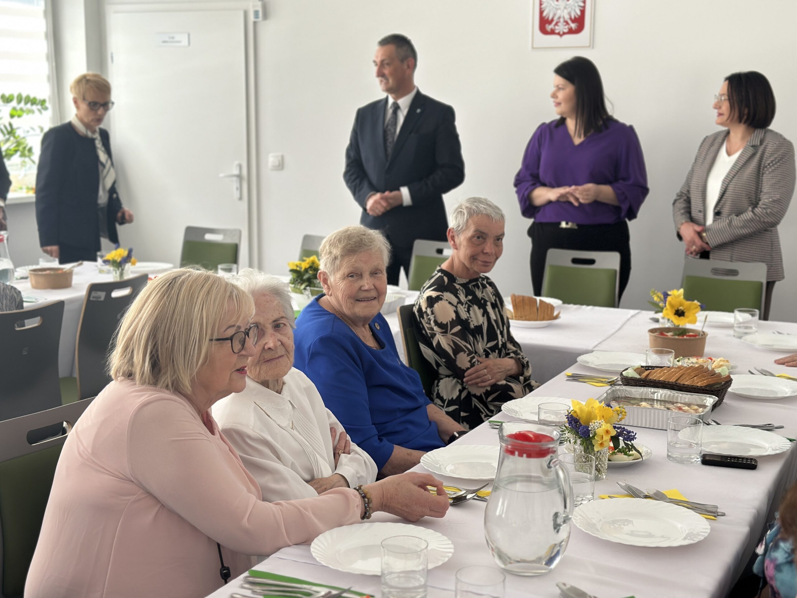 Na zdjęciu seniorki siedzące przy zastawionym stole. W tle widoczny wójt gminy, przewodnicząca rady gminy, skarbnik gminy, a także dyrektor GOPSu.