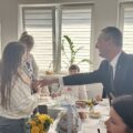 Na zdjęciu wójt wita się z dziewczynką. Dziewczynkę trzyma na rękach opiekunka ze żłobka. Na fotografii widać także seniorów.