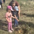 Na zdjęciu kobieta z dwójką dzieci - chłopcem i dziewczynką. Dzieci sadzą drzewko, a kobieta im pomaga.