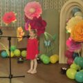 Na zdjęciu dziewczynka podczas swojego wystąpienia. W tle wystrój sceny - kolorowe duże kwiaty i balony.