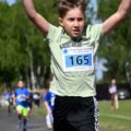 Zdjęcie przedstawia chłopca, który ma ręce uniesione w górze po przebiegnięciu trasy. W tle inni uczestniczy biegu. Chłopiec na koszulce ma przypiętą kartkę z numerem 165.