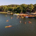 Zdjęcie wykonano "z lotu ptaka". Przedstawia osoby pływające kajakami po jeziorze. Na zdjęciu widać także plażę, drzewa i inną roślinność.